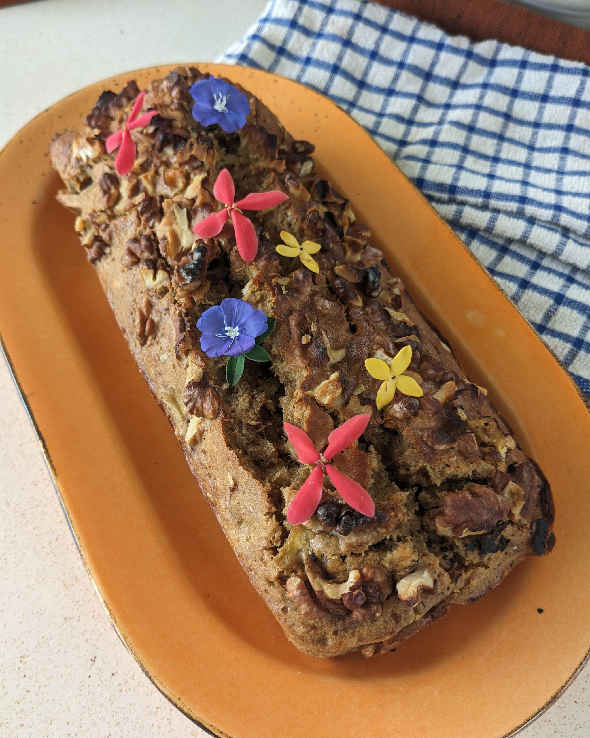 Banana bread disajikan diatas piring dengan topping kacang dan hiasan bungan diatasnya