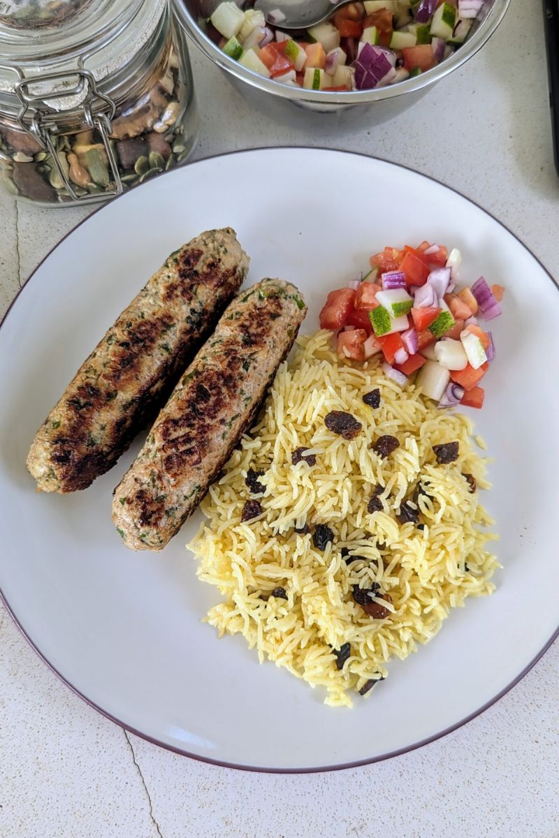 Sepiring hidangan berisi dua kebab panggang berbentuk sosis, nasi kuning dengan kismis, dan salad potongan tomat, bawang, serta timun