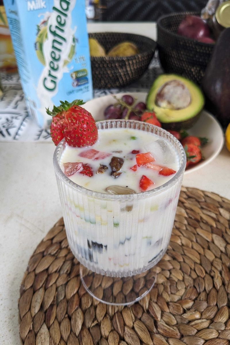 Minuman salad buah dingin dalam gelas bening dengan potongan stroberi di atasnya, disajikan di meja bersama buah-buahan segar dan susu Greenfields di latar belakang